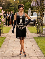 A woman stands on a stone pathway in a garden setting, wearing a black 1920s-style flapper dress with gold embroidery, long black gloves, and a black headband.
