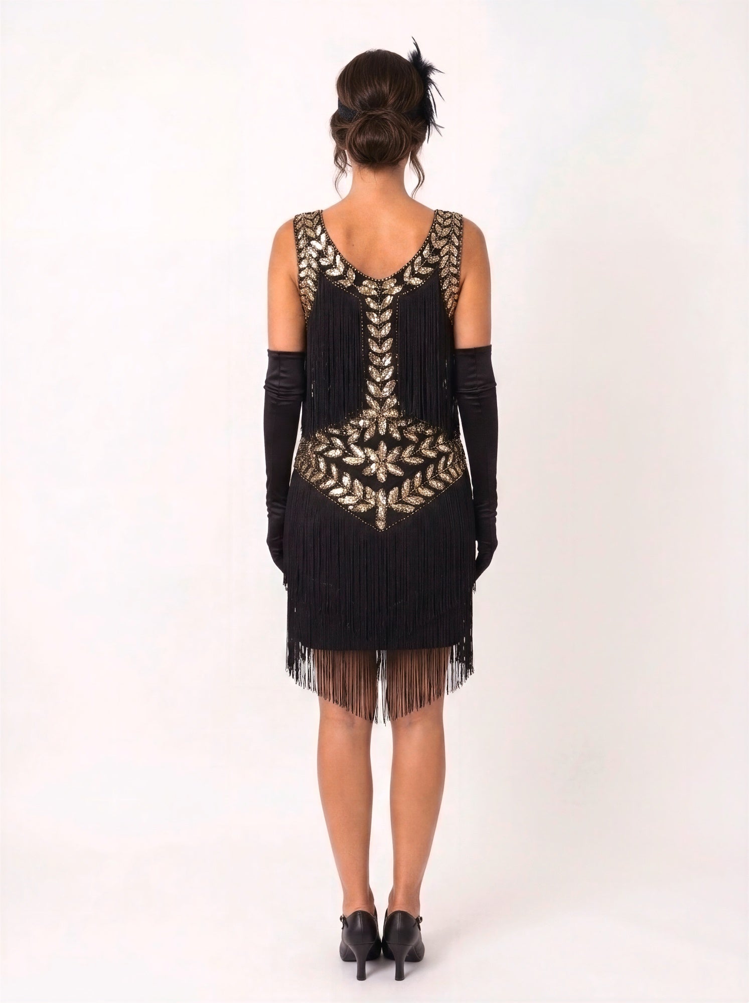 A woman viewed from the back wearing a black sleeveless fringe dress with ornate gold leaf embroidery across the neckline and back, paired with long black gloves and black heels, against a plain white studio background.