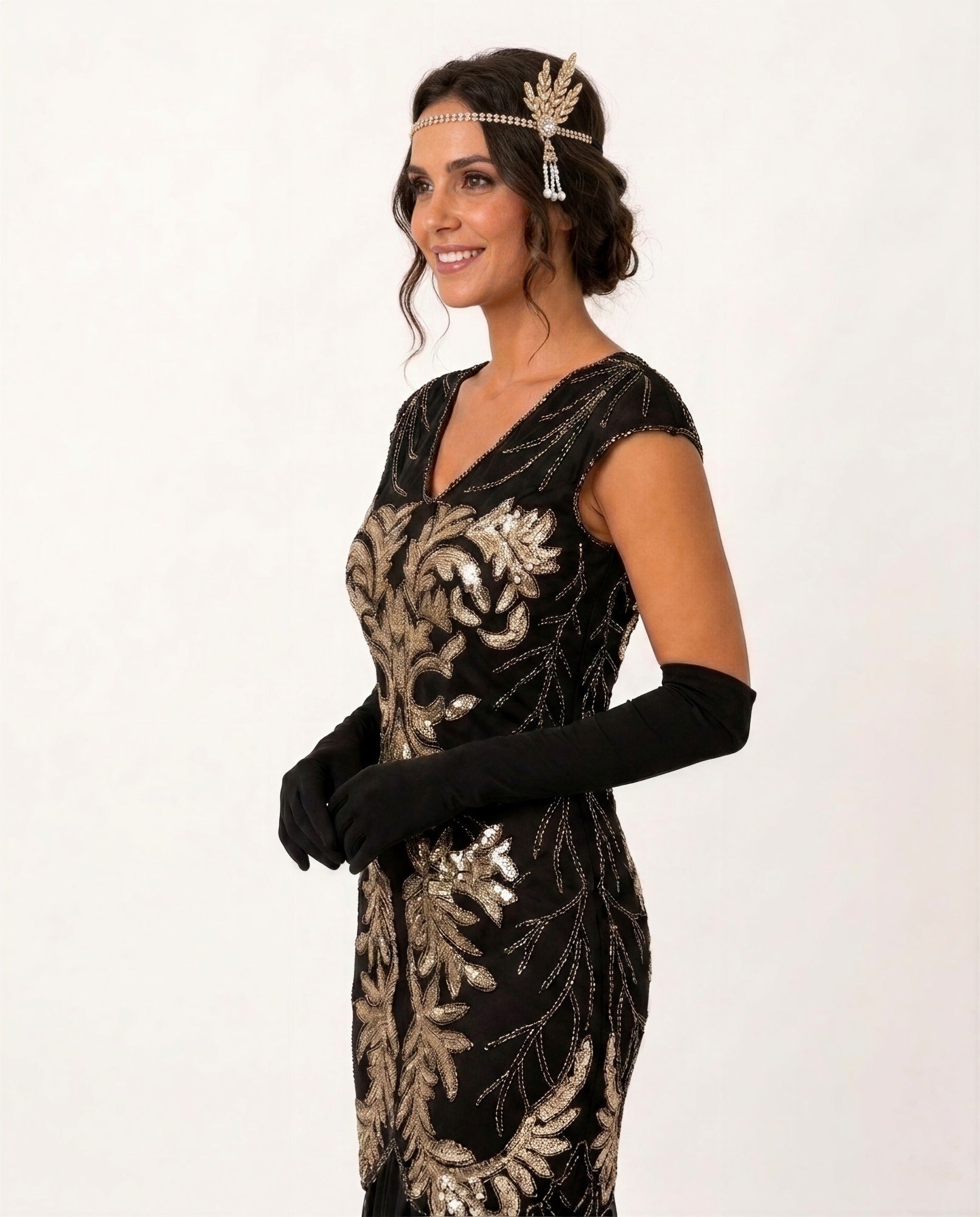 A woman modeling a 1920s-style flapper dress in black with ornate gold sequin embroidery, accessorized with long black gloves and a gold headband against a plain white background.