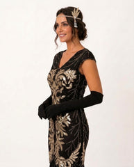 A woman modeling a 1920s-style flapper dress in black with ornate gold sequin embroidery, accessorized with long black gloves and a gold headband against a plain white background.
