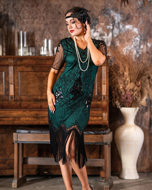 A woman models a 1920s-style green flapper dress with black fringe and sequins, accessorized with a pearl necklace and headband, posed in front of a vintage wooden piano.
