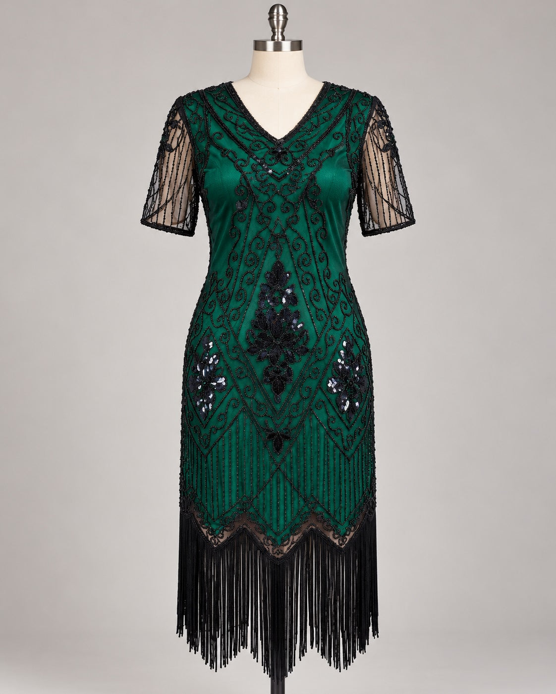 A dark green 1920s-style flapper dress displayed on a mannequin against a neutral gray background. The dress features black sequin and bead geometric patterns, sheer black mesh short sleeves, and a long black fringe hem.