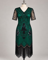 A dark green 1920s-style flapper dress displayed on a mannequin against a neutral gray background. The dress features black sequin and bead geometric patterns, sheer black mesh short sleeves, and a long black fringe hem.
