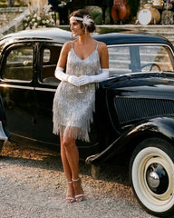 A woman in a 1920s-style silver fringe flapper dress, white opera gloves, and a matching headband stands beside a vintage black car outdoors.