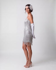 A woman modeling a silver 1920s-style flapper dress with fringe, white opera gloves, a feathered headband, and clear high-heeled sandals against a light gray studio background.