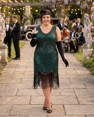 A woman in a 1920s-style green plus size flapper dress with black fringe, long black gloves, and a headband walks down a stone path at an outdoor event, holding a glass of white wine.