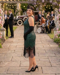 A woman stands on a stone pathway at an outdoor event, wearing a dark green 1920s-style plus size flapper dress with black fringe, long black gloves, and a black headpiece. Guests in tuxedos and a band are visible in the background.
