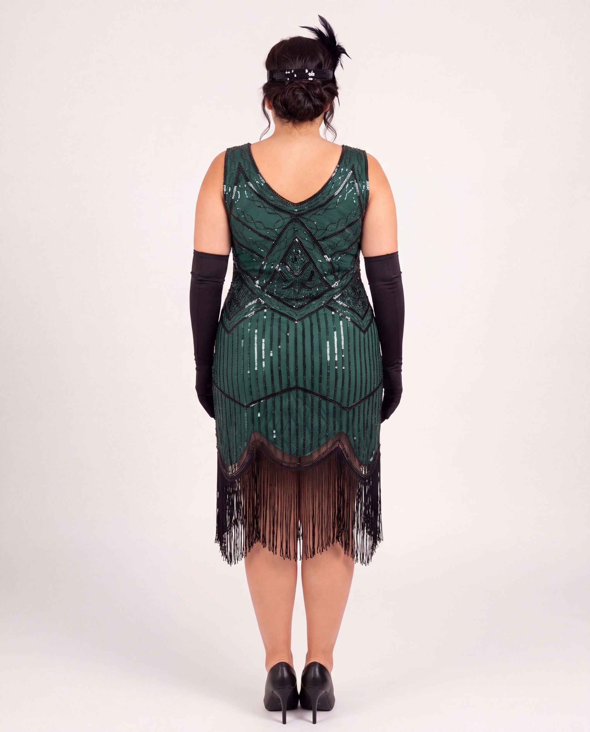 A woman shown from the back wearing a dark green 1920s-style plus size flapper dress with black fringe, black elbow-length gloves, and black heels against a plain light gray studio background.