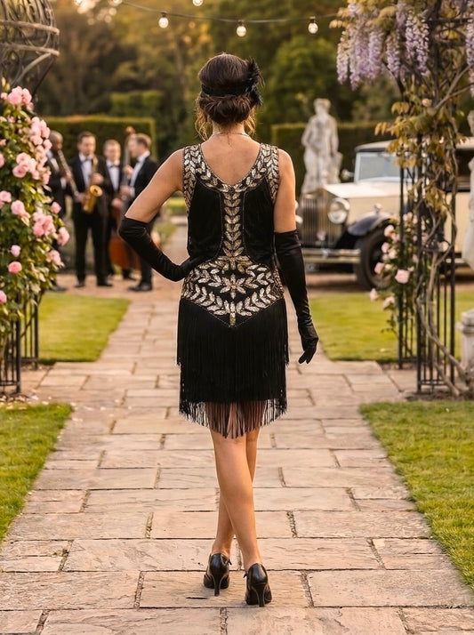 A woman viewed from behind wearing a black 1920s-style flapper dress with gold beaded geometric patterns and long black gloves, walking down a stone path in a garden setting.