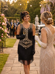 A woman in a 1920s-style black flapper dress with gold embroidery and fringe, holding a glass of champagne at an outdoor garden party, standing next to another woman in a sparkling light-colored gown.