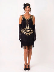 A woman modeling a black 1920s-style flapper dress with gold leaf embroidery, black fringe, long black gloves, a black headband, and black heels against a plain white studio background.