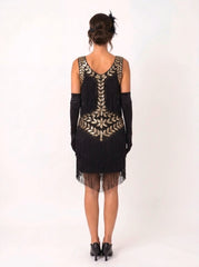 A woman viewed from the back wearing a black sleeveless fringe dress with ornate gold leaf embroidery across the neckline and back, paired with long black gloves and black heels, against a plain white studio background.