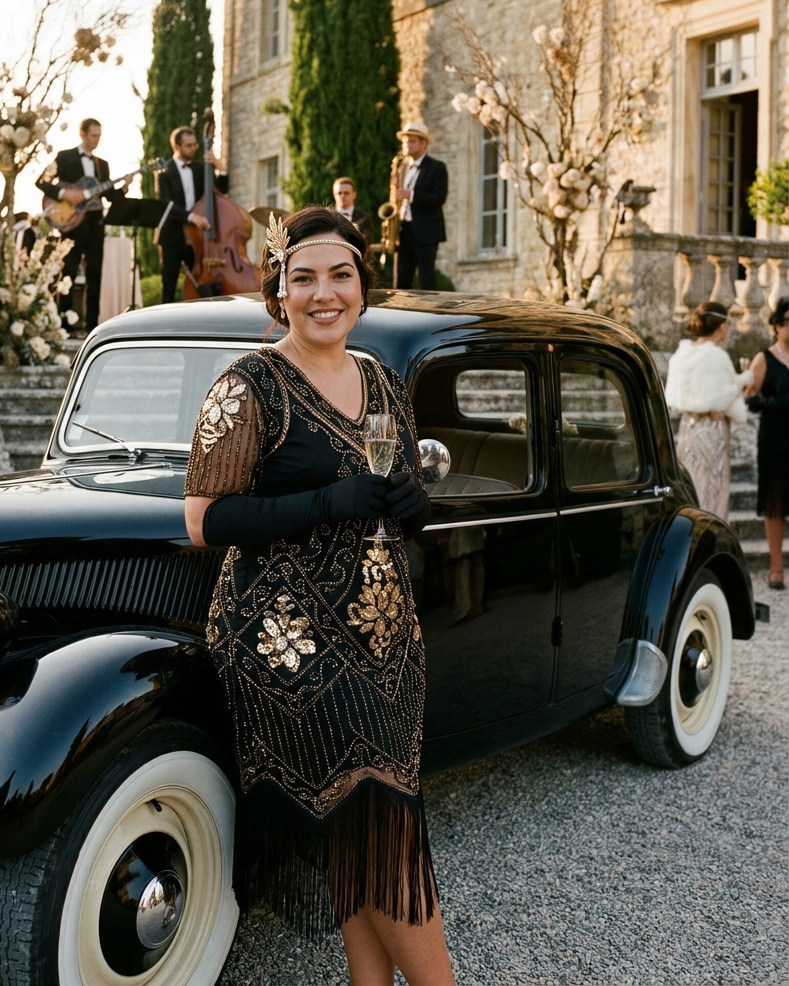 A woman in a 1920s-style flapper dress and headpiece stands in front of a vintage black car at an outdoor event, holding a glass. The dress is black with ornate gold and beige floral embroidery and fringe details.