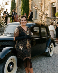 A woman in a 1920s-style flapper dress and headpiece stands in front of a vintage black car at an outdoor event, holding a glass. The dress is black with ornate gold and beige floral embroidery and fringe details.