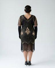 A woman viewed from the back wearing a black 1920s-style flapper dress with gold embroidery, fringe, and sheer short sleeves, paired with long black gloves and black heels, against a light gray studio background.