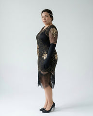 A woman modeling a black 1920s-style flapper dress with gold sequin embroidery, sheer long sleeves, and a fringe hem, paired with black gloves and heels against a light gray studio background.