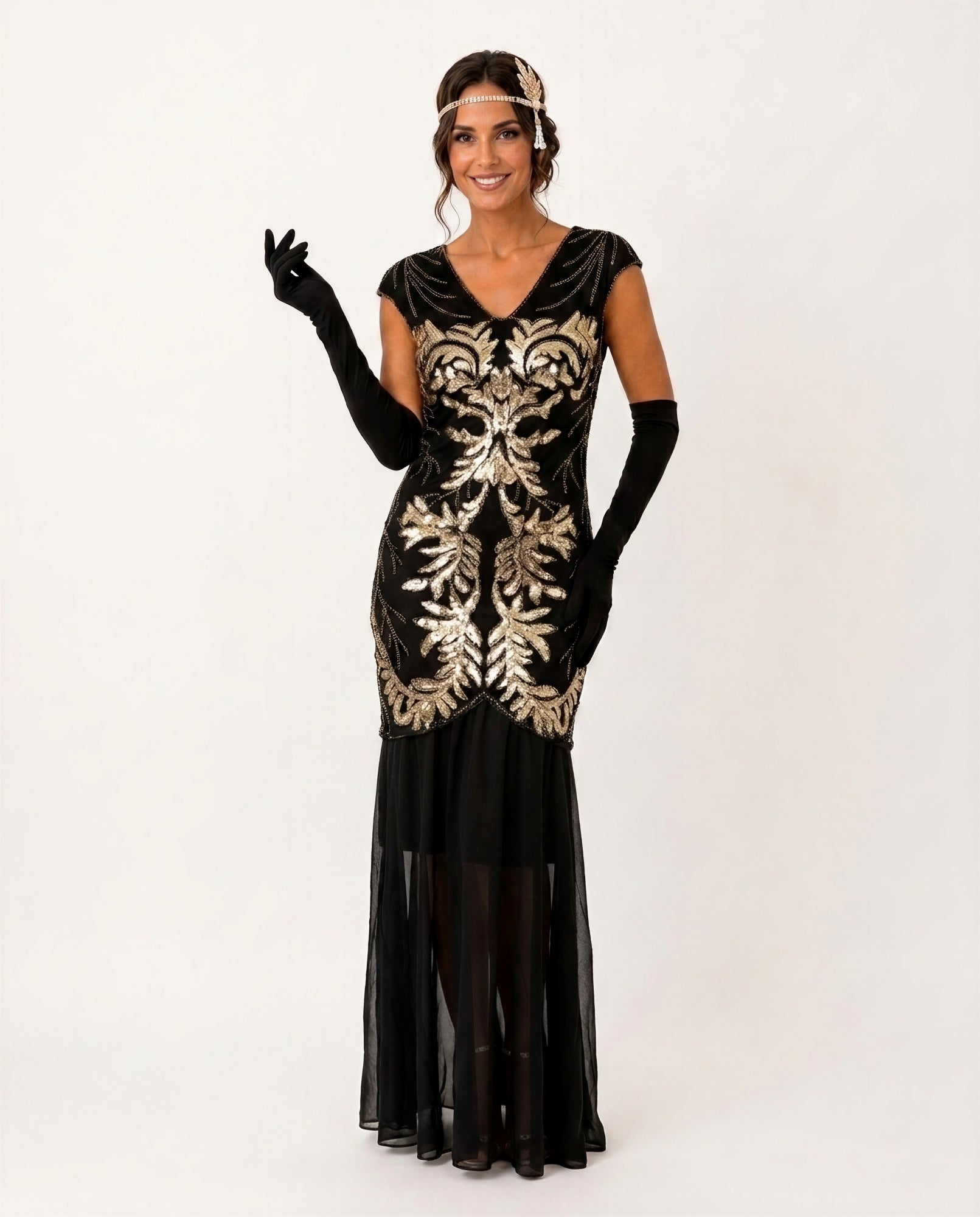 A woman modeling a 1920s-style flapper dress in black velvet with ornate gold sequin embroidery, accessorized with long black gloves, a headband, and sheer black stockings against a plain white studio background.