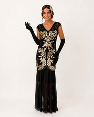 A woman modeling a 1920s-style flapper dress in black velvet with ornate gold sequin embroidery, accessorized with long black gloves, a headband, and sheer black stockings against a plain white studio background.