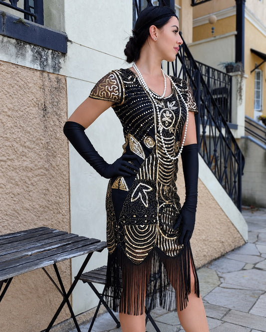 A woman modeling a 1920s-style black flapper dress with gold geometric beadwork and fringe, accessorized with long black gloves and pearl necklaces, standing outdoors against a building wall.