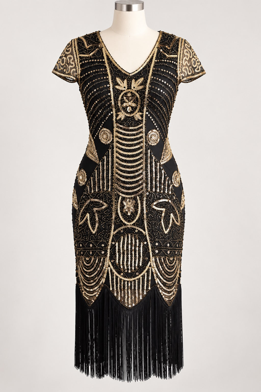 A black 1920s-style flapper dress with elaborate gold beaded and sequined Art Deco patterns, short sleeves, a V-neckline, and a long black fringe hem, displayed on a mannequin against a light gray background.