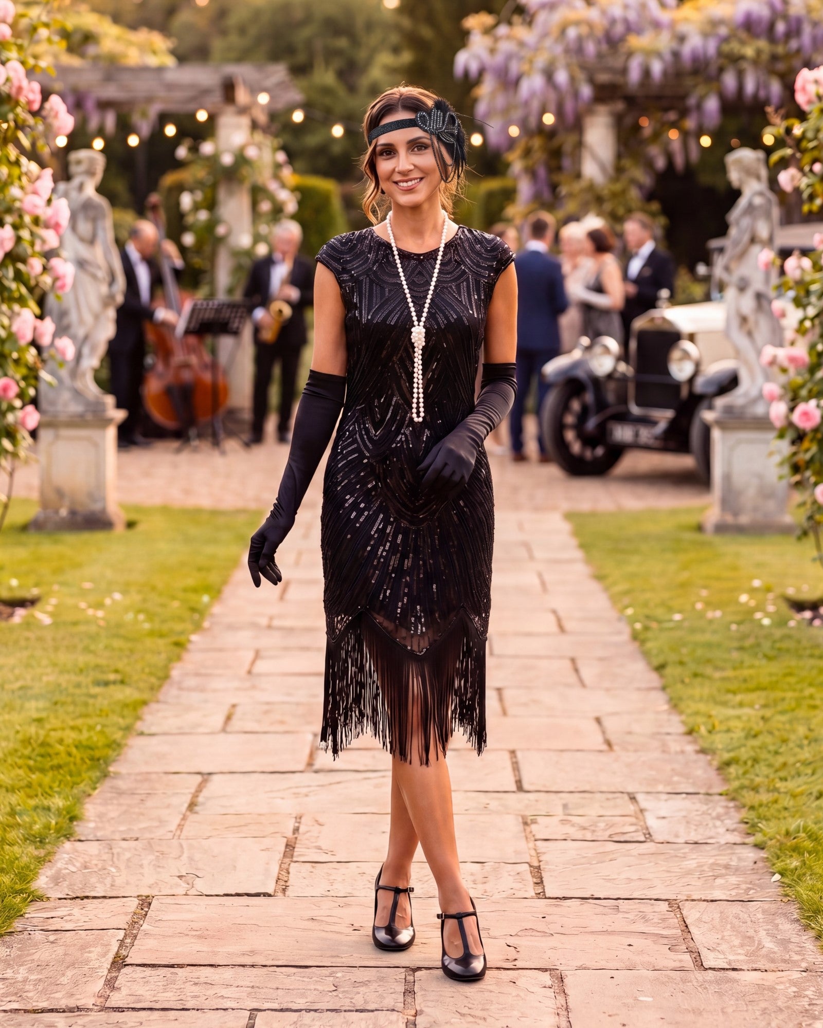 A woman stands on a stone path in a garden wearing a black 1920s-style flapper dress with fringe, long black gloves, a black headband, and a long silver necklace. Guests and a vintage car are blurred in the background.