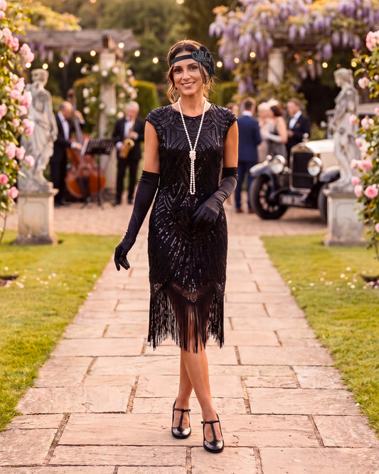 A woman stands on a stone path in a garden wearing a black 1920s-style flapper dress with fringe, long black gloves, a black headband, and a long silver necklace. Guests and a vintage car are blurred in the background.