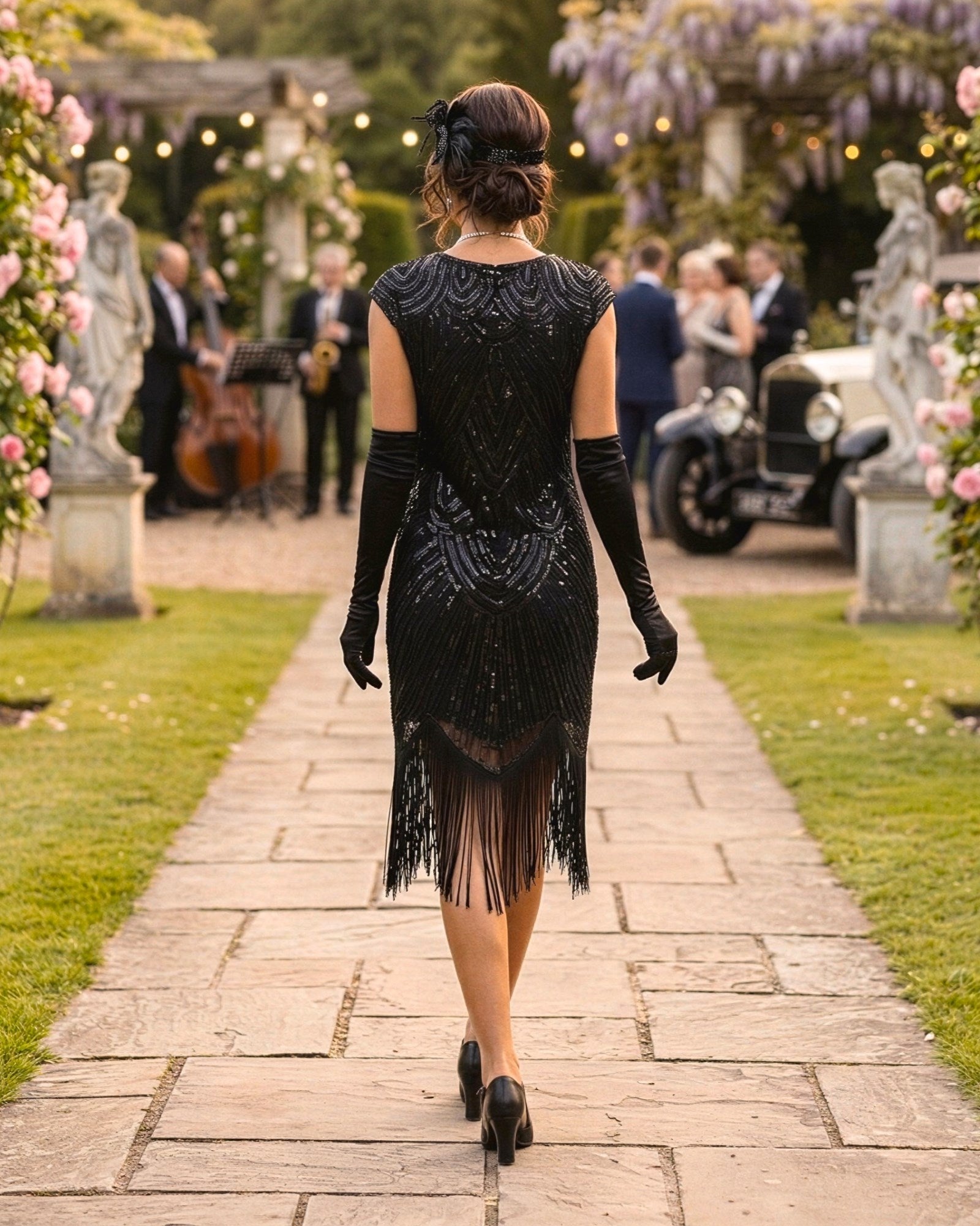 A woman viewed from behind walking down a stone path in a garden, wearing a black sequined flapper-style dress with fringe, long black gloves, and black heels. Guests, flowers, statues, and an old car are visible in the background.