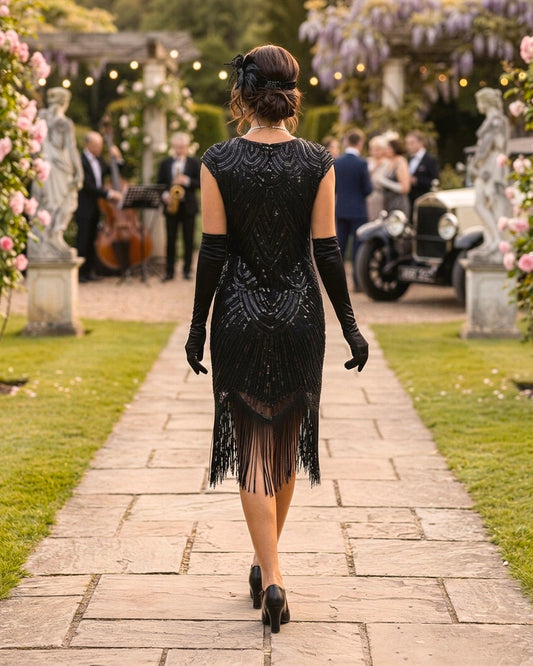 A woman viewed from behind walking down a stone path in a garden, wearing a black sequined flapper-style dress with fringe, long black gloves, and black heels. Guests, flowers, statues, and an old car are visible in the background.