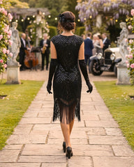 A woman viewed from behind walking down a stone path in a garden, wearing a black sequined flapper-style dress with fringe, long black gloves, and black heels. Guests, flowers, statues, and an old car are visible in the background.