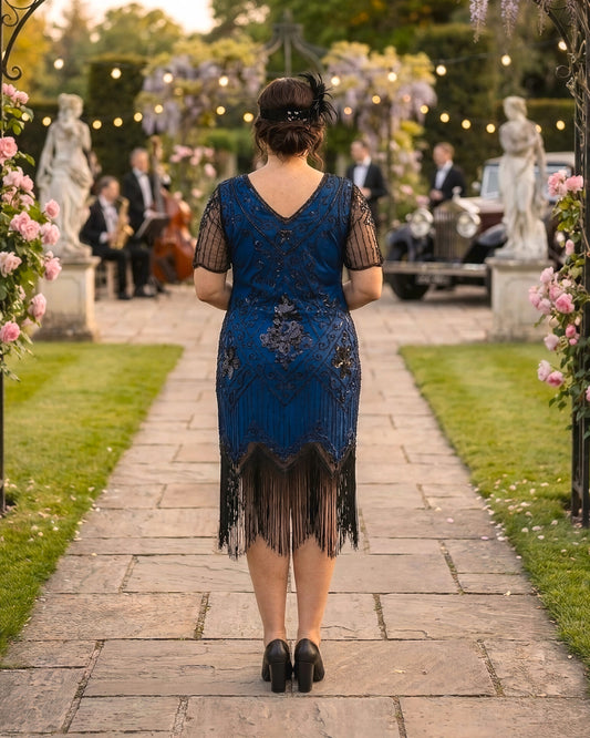 A woman viewed from behind wearing a blue 1920s-style flapper dress with black fringe and lace, standing on a stone path in a garden setting with guests and string lights in the background.