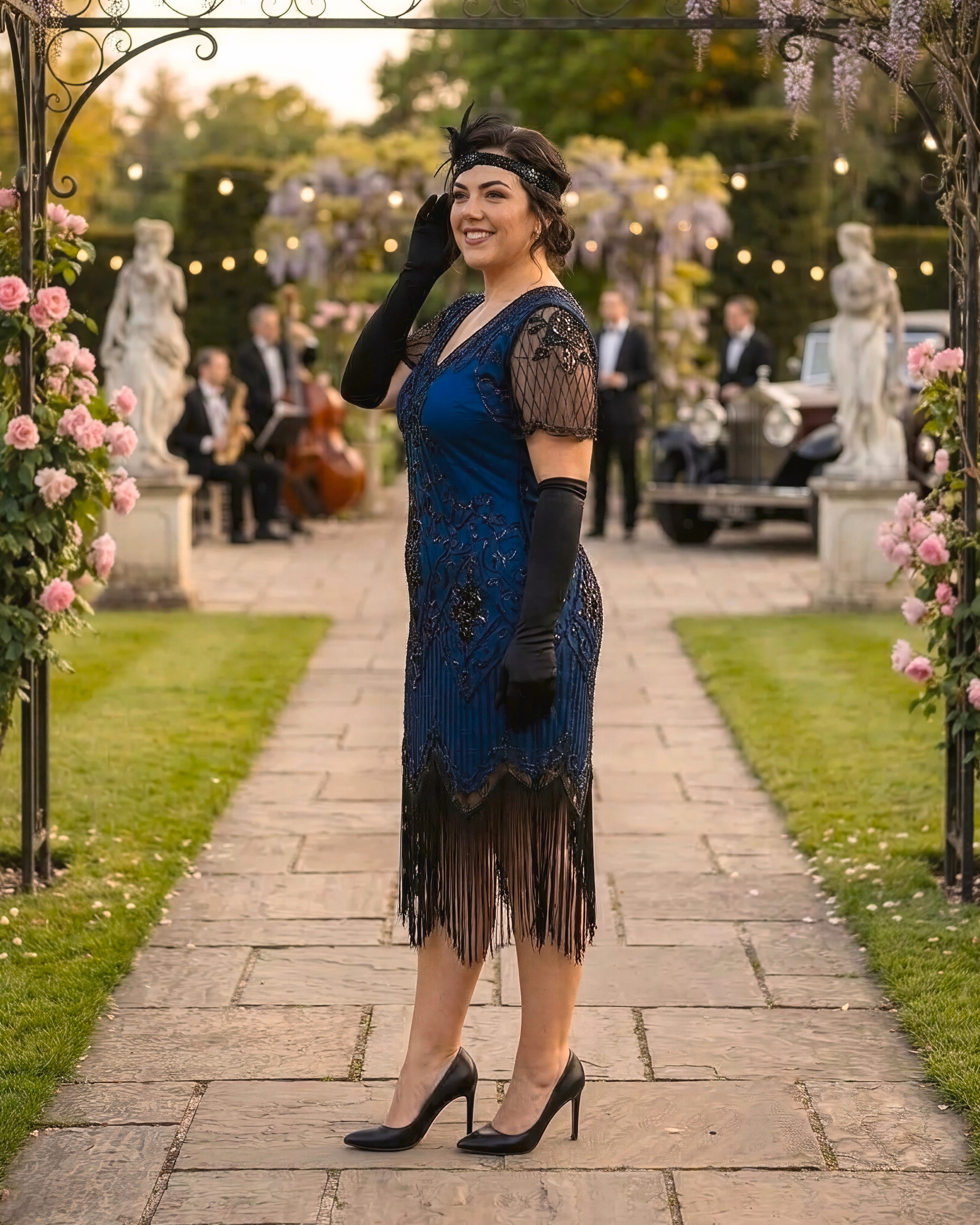A woman stands on a stone path in a garden wearing a blue and black 1920s-style flapper dress with black gloves, a black headband, and black heels. The background shows a softly blurred outdoor party scene with statues, flowers, lights, and musicians.
