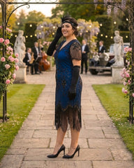 A woman stands on a stone path in a garden wearing a blue and black 1920s-style flapper dress with black gloves, a black headband, and black heels. The background shows a softly blurred outdoor party scene with statues, flowers, lights, and musicians.