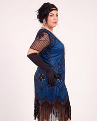 A woman modeling a blue 1920s-style flapper dress with black fringe, black mesh sleeves, and black elbow-length gloves against a plain light background.