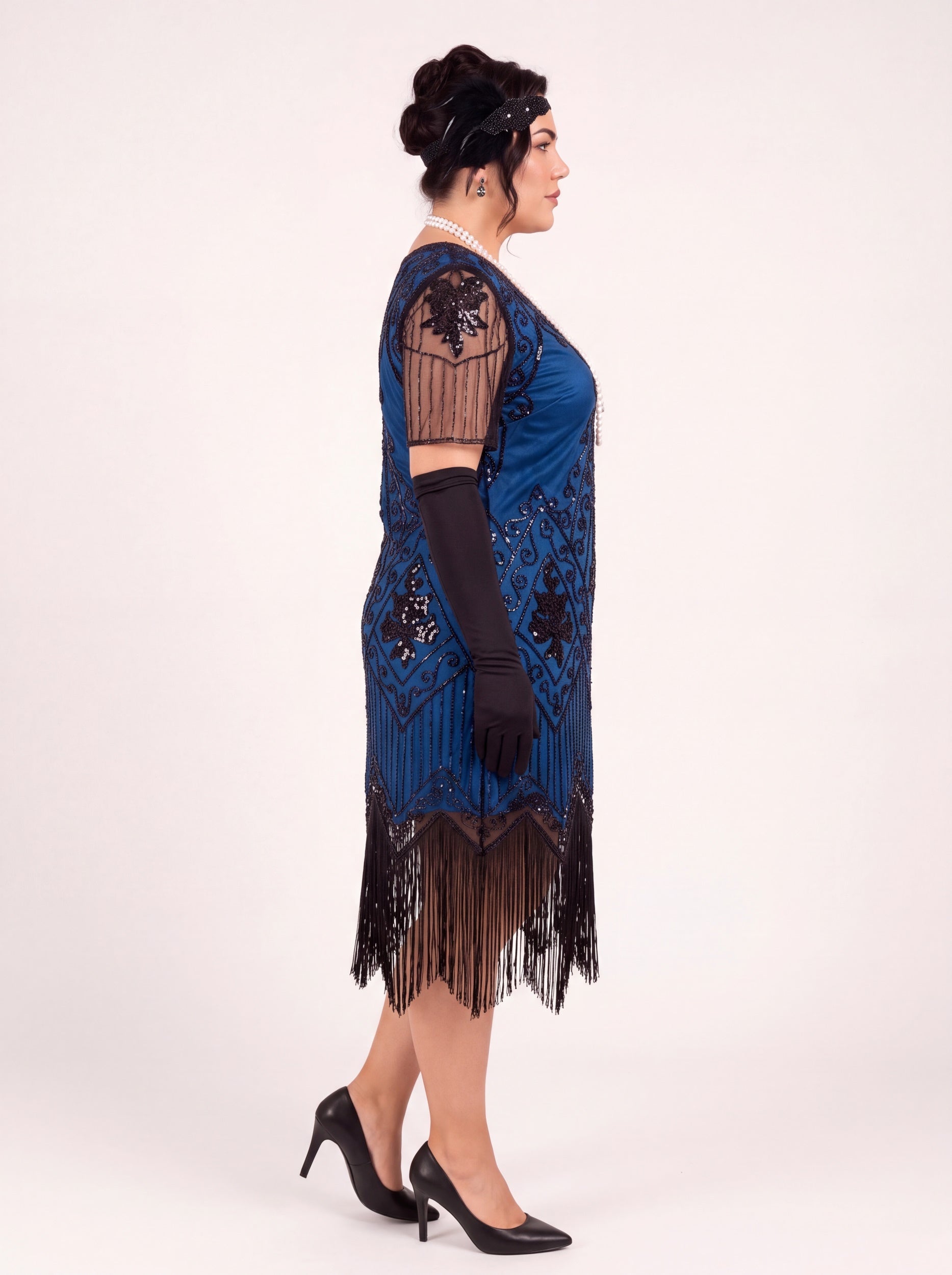 A woman modeling a blue 1920s-style flapper dress with black fringe, black lace sleeves, black gloves, and black heels against a plain light background.