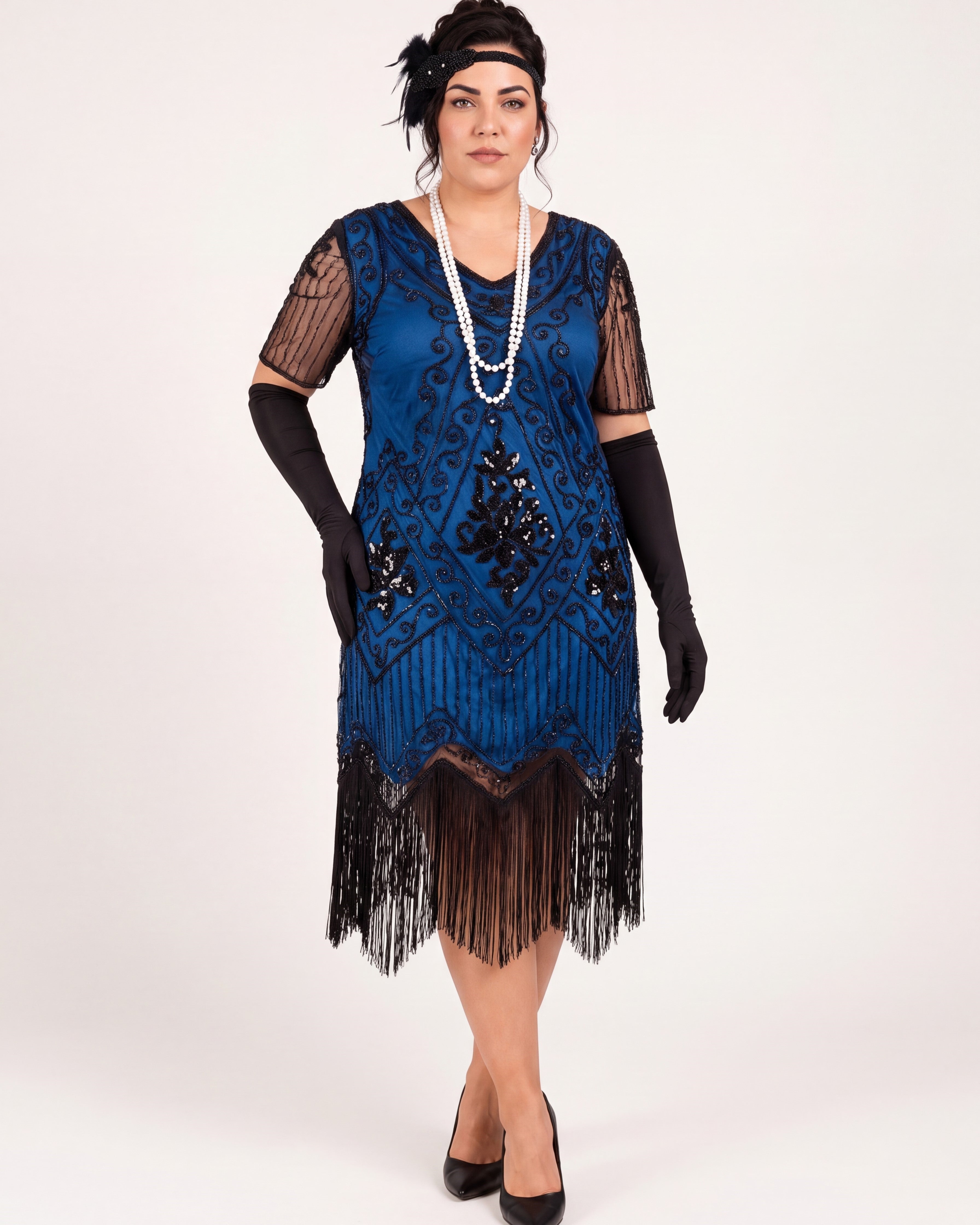 A woman modeling a blue 1920s-style flapper dress with black fringe, sheer black mesh sleeves, black gloves, a long pearl necklace, and a black headpiece against a plain light background.