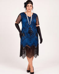 A woman modeling a blue 1920s-style flapper dress with black fringe, sheer black mesh sleeves, black gloves, a long pearl necklace, and a black headpiece against a plain light background.
