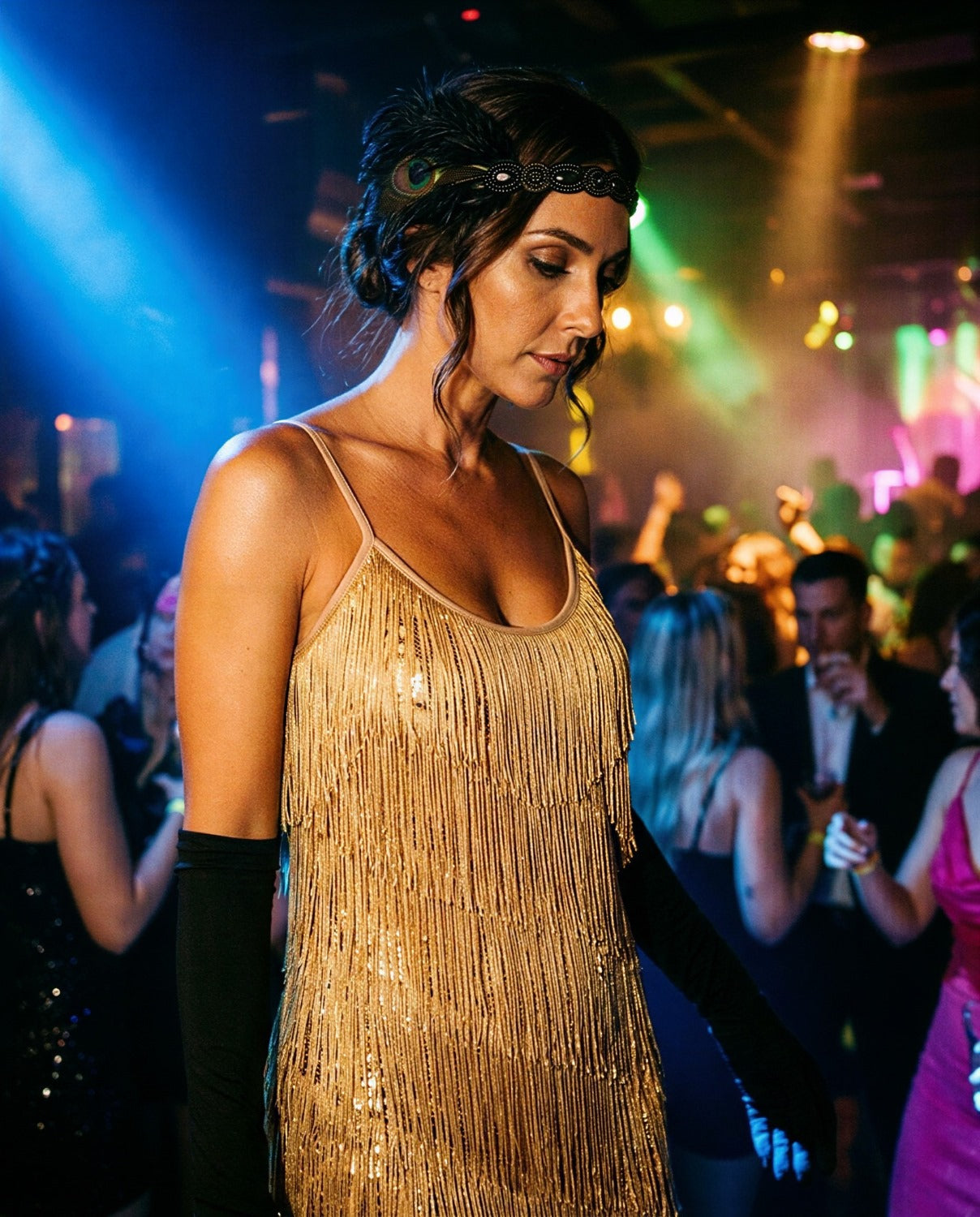 A woman at a nightclub or party wearing a gold fringe & sequin 1920s flapper dress with black elbow-length gloves and a black feathered headband. The background shows a dimly lit dance floor with colored stage lighting and other people.
