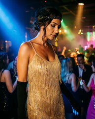 A woman at a nightclub or party wearing a gold fringe & sequin 1920s flapper dress with black elbow-length gloves and a black feathered headband. The background shows a dimly lit dance floor with colored stage lighting and other people.