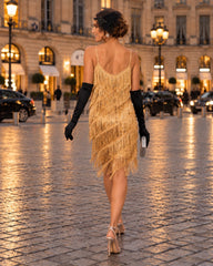 A woman wearing a gold fringe & sequin 1920s flapper dress, black elbow-length gloves, and beige high heels walks down a cobblestone street at dusk, viewed from behind.