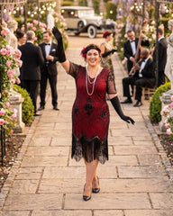 A plus size woman in a 1920s-style flapper dress walks down a stone pathway at a garden wedding or event. The dress is deep red with black fringe, black gloves, and black hair accessories, surrounded by guests, flowers, and string lights.