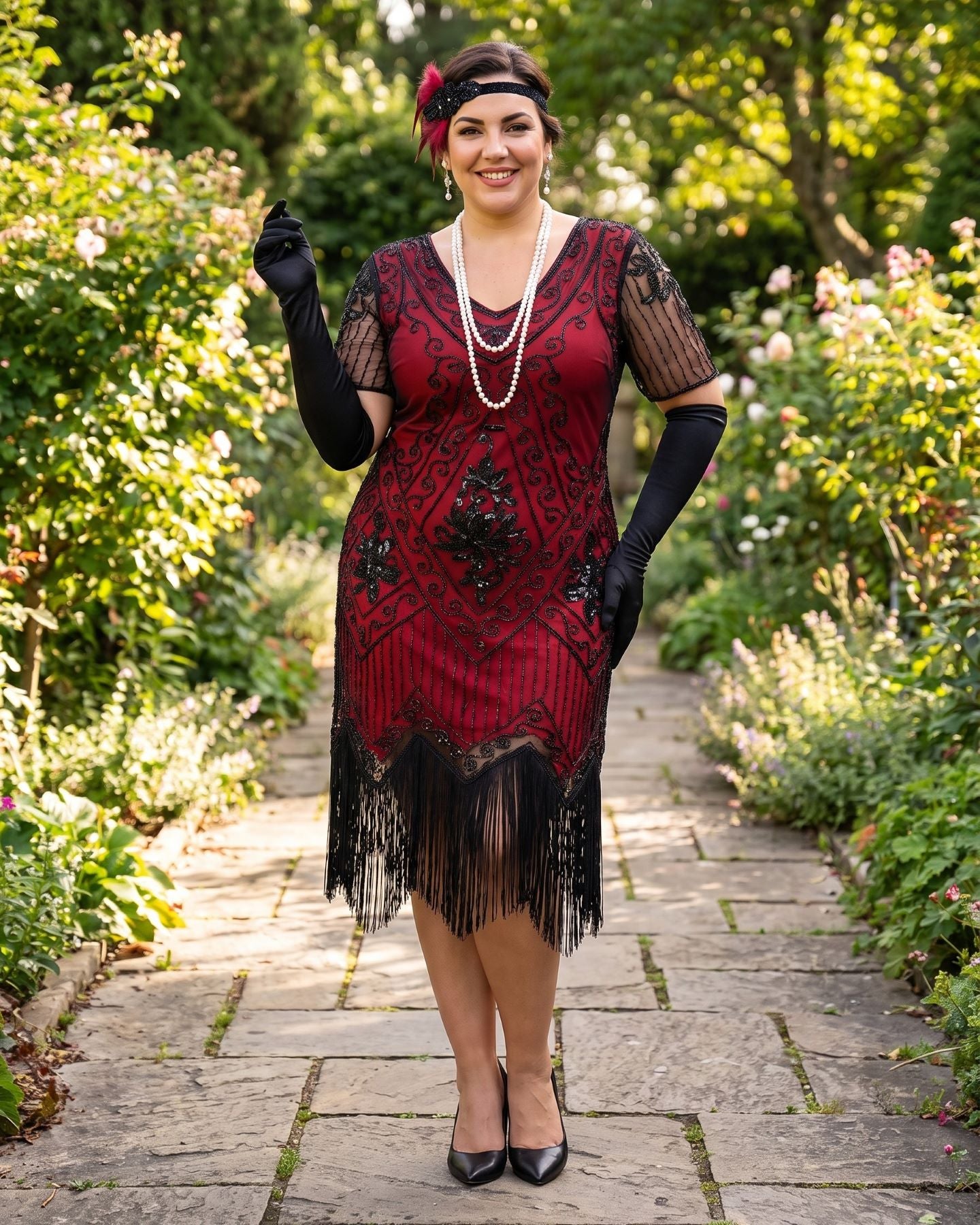 A plus size woman stands on a stone garden path wearing a 1920s flapper-style dress. The dress is deep red with black lace patterns and a black fringe hem.