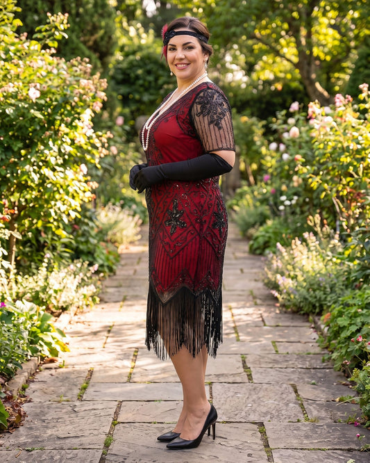 A plus size woman stands on a stone garden path wearing a red and black 1920s-style flapper dress with black gloves, a headband, and black heels, with lush green garden foliage in the background.