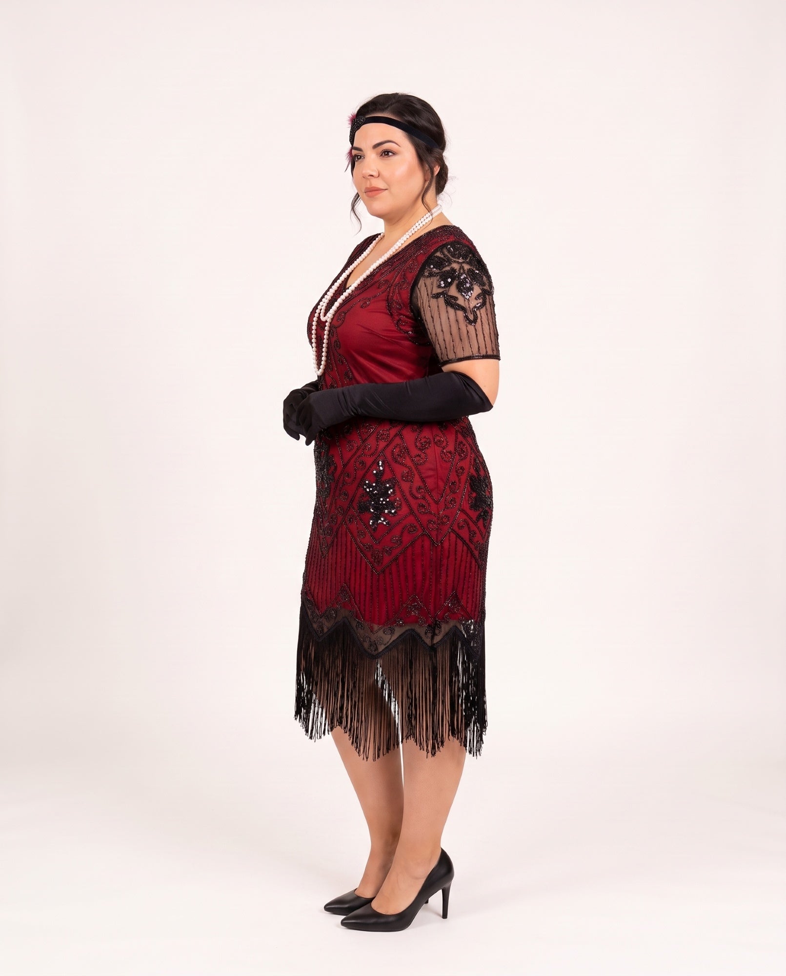 A woman modeling a plus size 1920s-style flapper dress in deep red and black, featuring fringe, lace sleeves, and black gloves, posed against a plain white studio background.