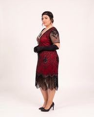 A woman modeling a plus size 1920s-style flapper dress in deep red and black, featuring fringe, lace sleeves, and black gloves, posed against a plain white studio background.