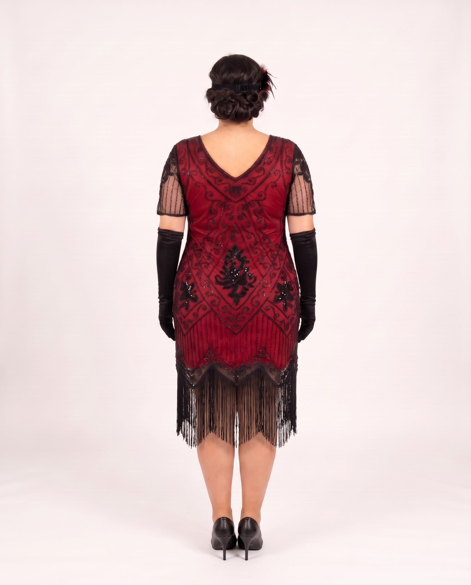 A woman viewed from the back wearing a red and black plus size 1920s-style flapper dress with black gloves and black heels, standing against a plain light background.