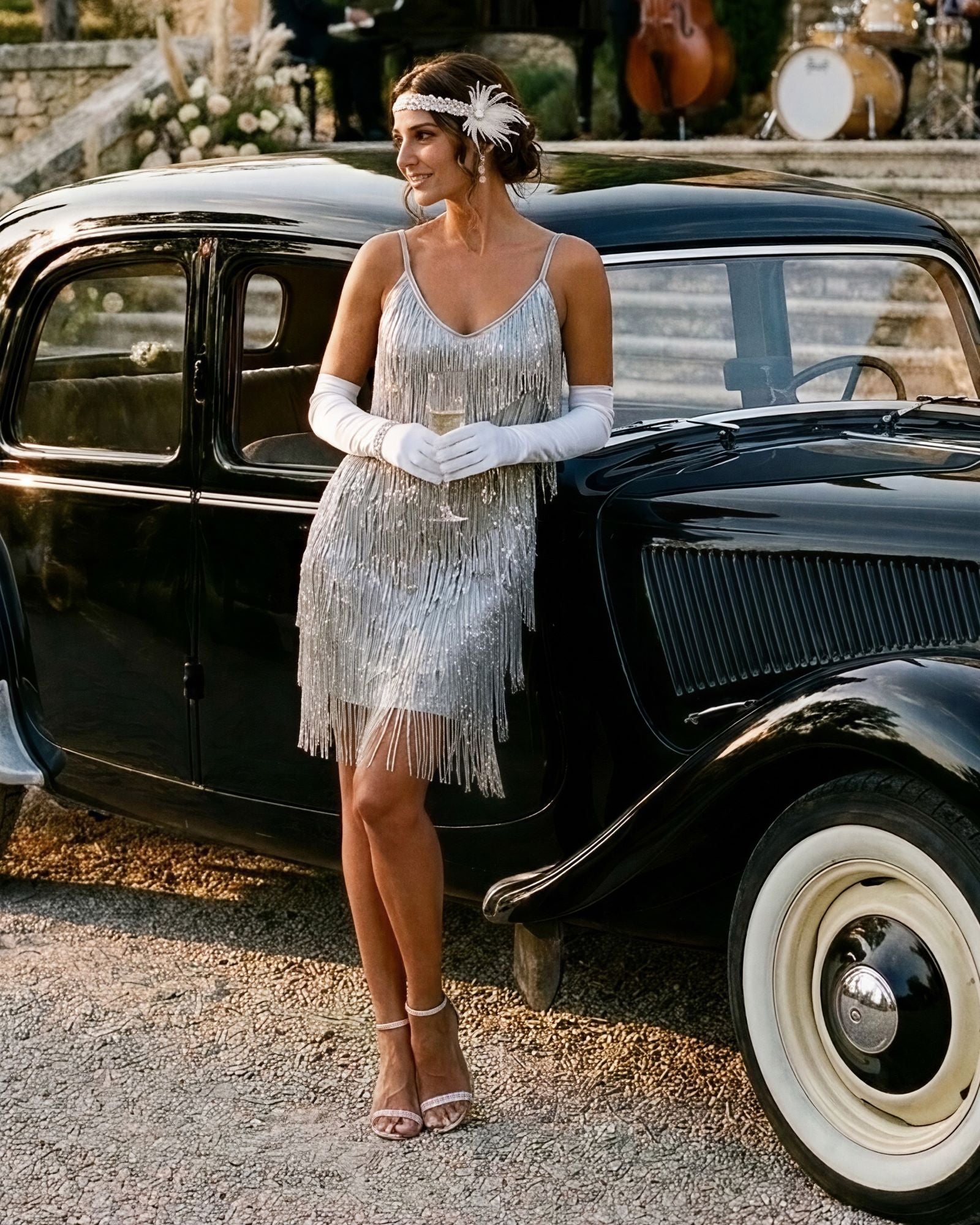 A woman in a 1920s-style silver fringe flapper dress, white opera gloves, and a matching headband stands beside a vintage black car outdoors.