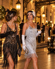 Two women in 1920s flapper-style dresses walking on a cobblestone street at night. One wears a black fringed dress with gold embellishments, long black gloves, and a feather headpiece.