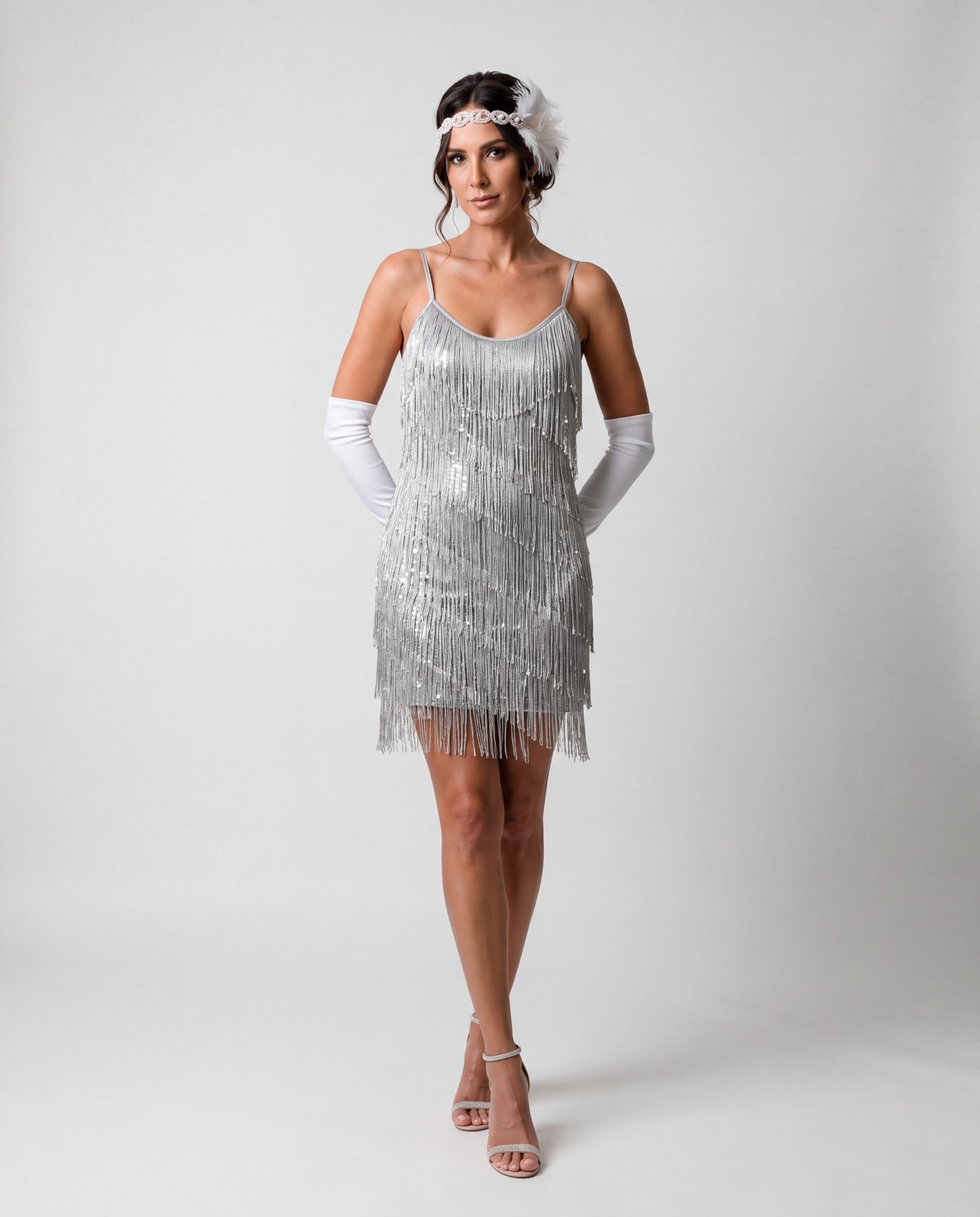 A woman modeling a silver 1920s-style flapper dress with fringe, white opera gloves, a white headband, and clear high-heeled sandals against a light gray studio background.