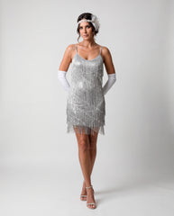 A woman modeling a silver 1920s-style flapper dress with fringe, white opera gloves, a white headband, and clear high-heeled sandals against a light gray studio background.