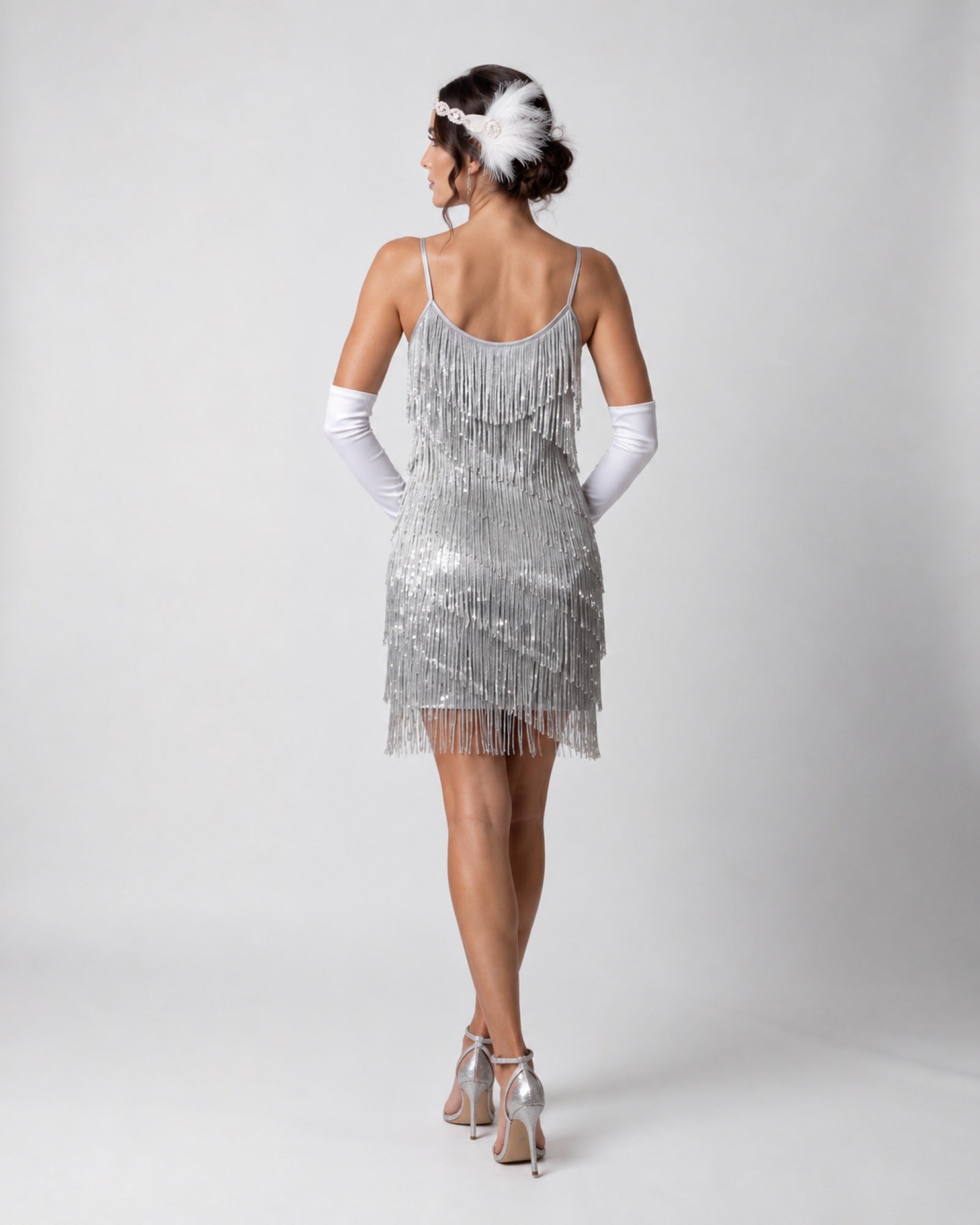 A woman shown from the back wearing a silver sequined fringe dress, white opera gloves, and silver high heels against a light gray studio background.